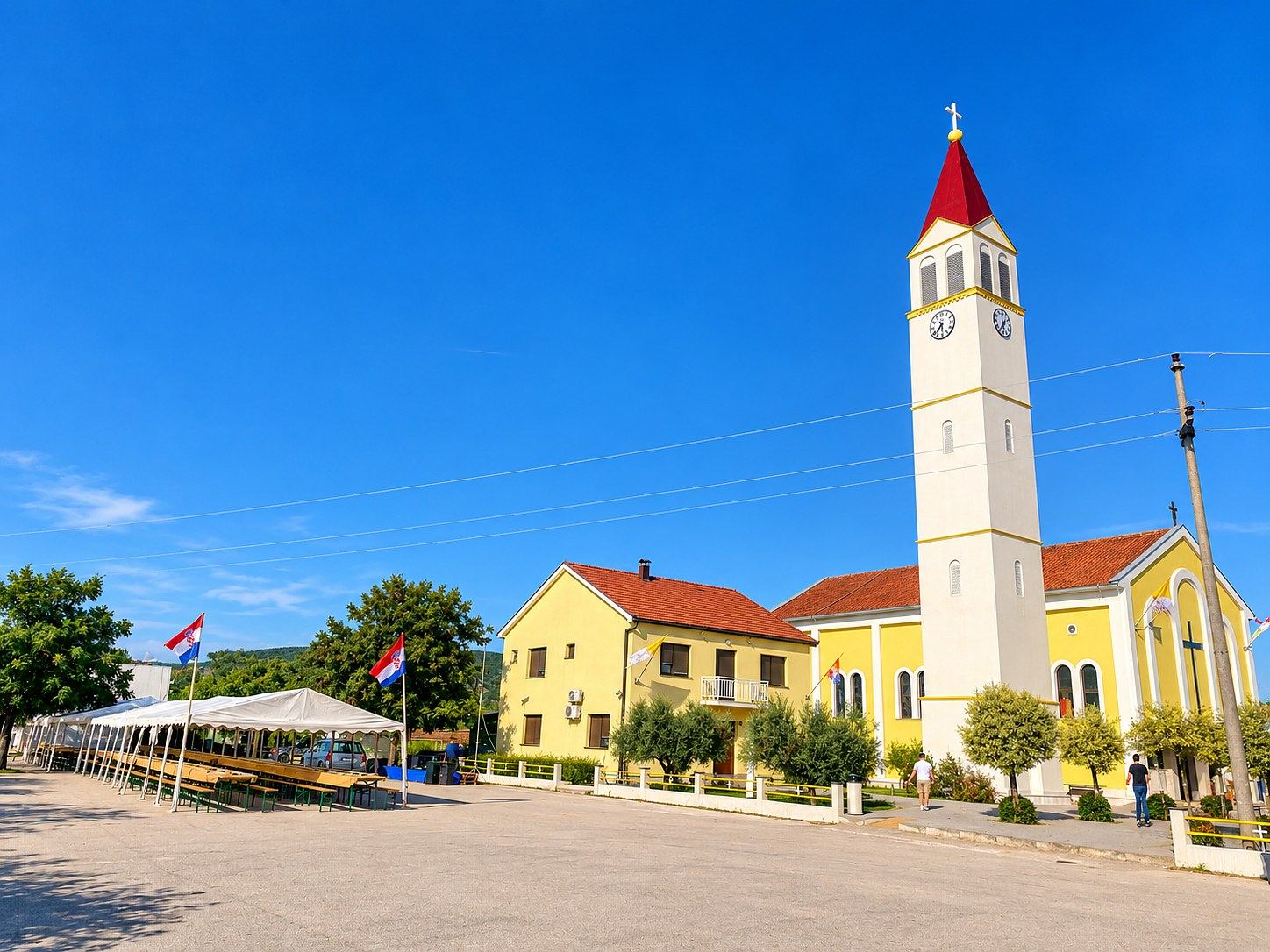 The yellow Church of the Sacred Heart of Jesus in Čeljevo with its tall white bell tower and red spire, beside the parish house