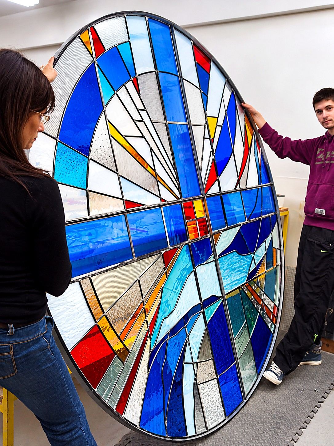 Round stained glass window by Brenda Belfield for Heritage Fellowship Church, Reston, Virginia, held upright by studio assistant Milan Stanisic Junior in the Stanisic Studio before delivery: a cross with rays of gold and red over flowing blue glass