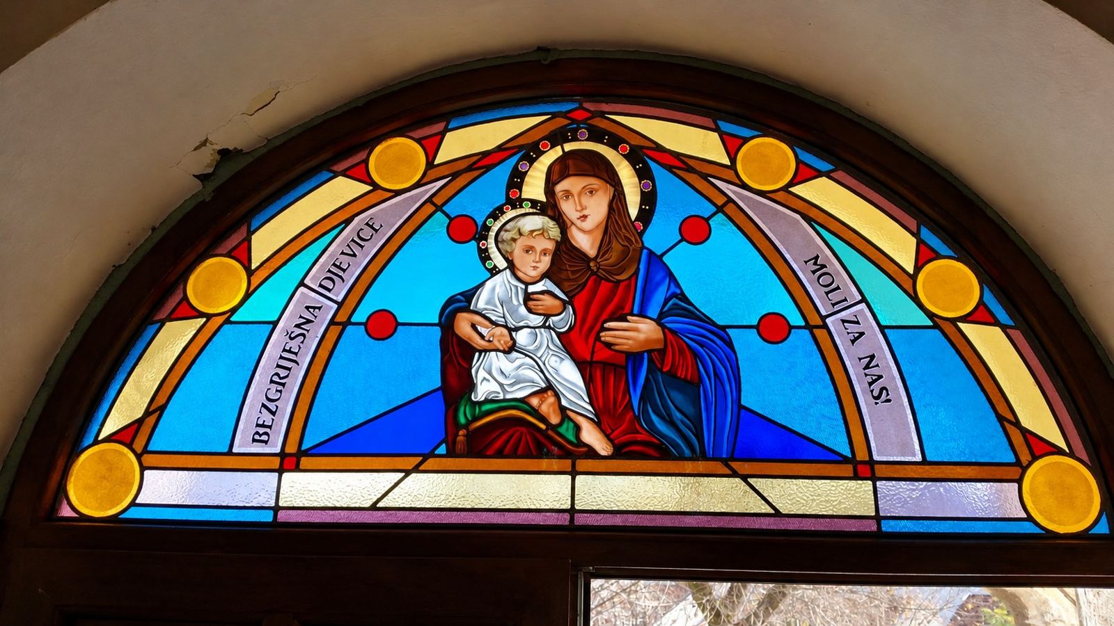 Stained glass lunette of Mary with the Child and the words Bezgrješna Djevice moli za nas above the door of the church in Hrasno