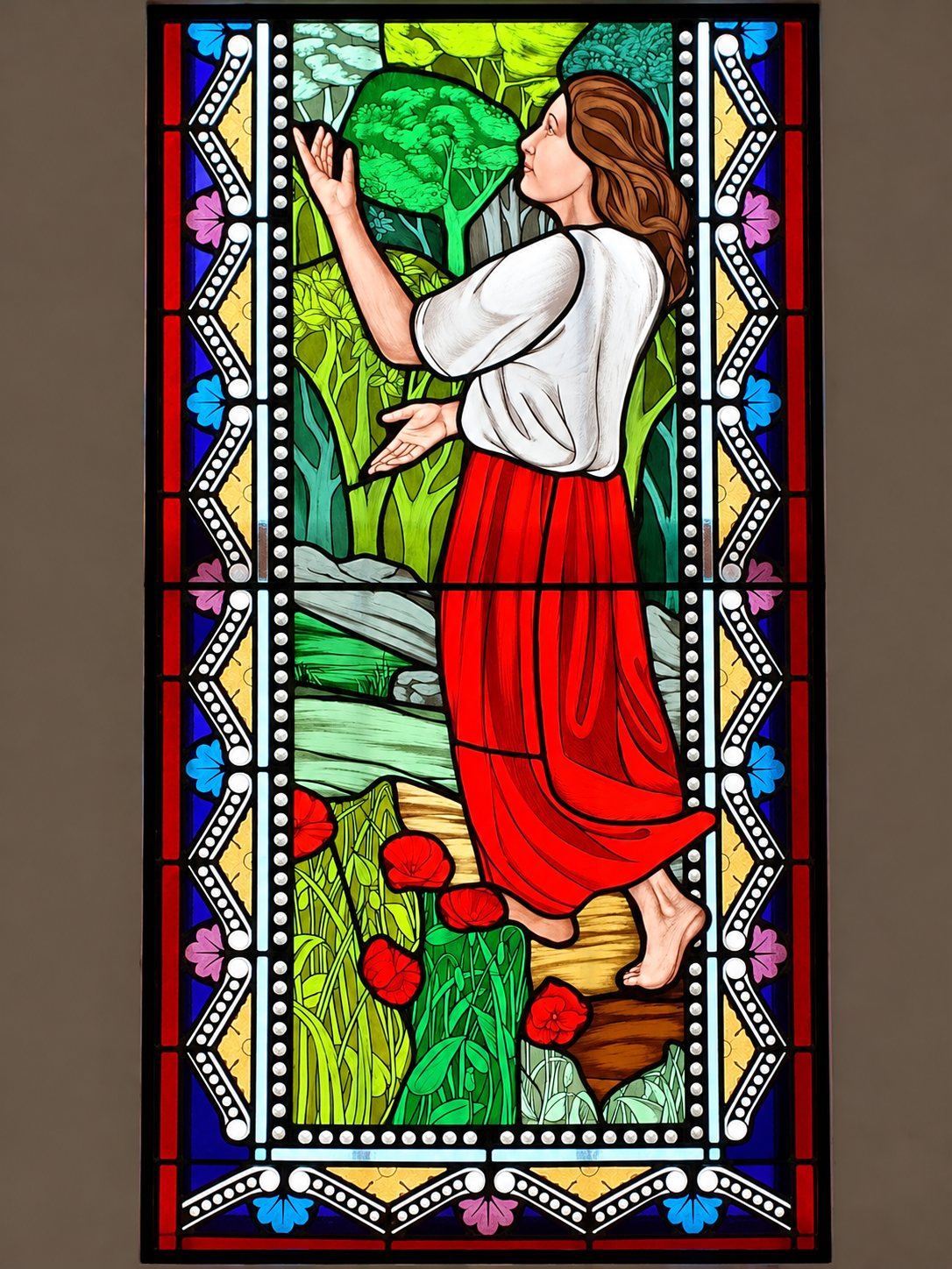 Stained glass window of a girl in a red skirt reaching up among the trees, Pastoral Centre in Tuzi