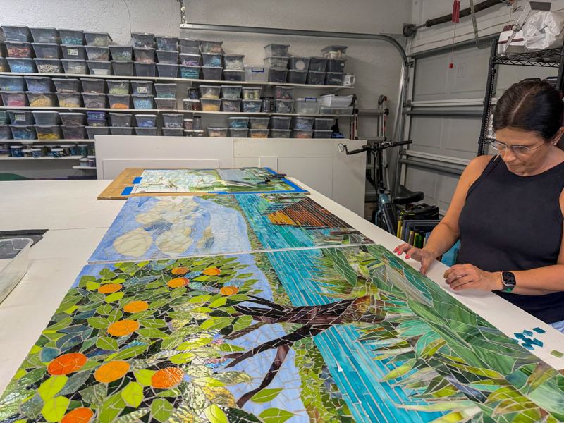 Three connected glass mosaic panels forming a Florida lake triptych, with a citrus tree on the left, a wooden dock under a cloudy sky in the center, and a great egret in flight over a heron in the marsh on the right, displayed in the Apperlo Art studio in Bonita Springs.