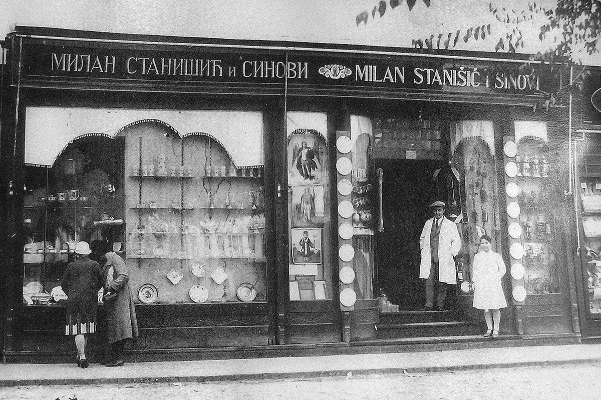 Стара црно бела фотографија излога породичне радње, са натписом Милан Станишић и синови исписаним ћирилицом и латиницом, и стаклом, порцеланом и иконама у излозима