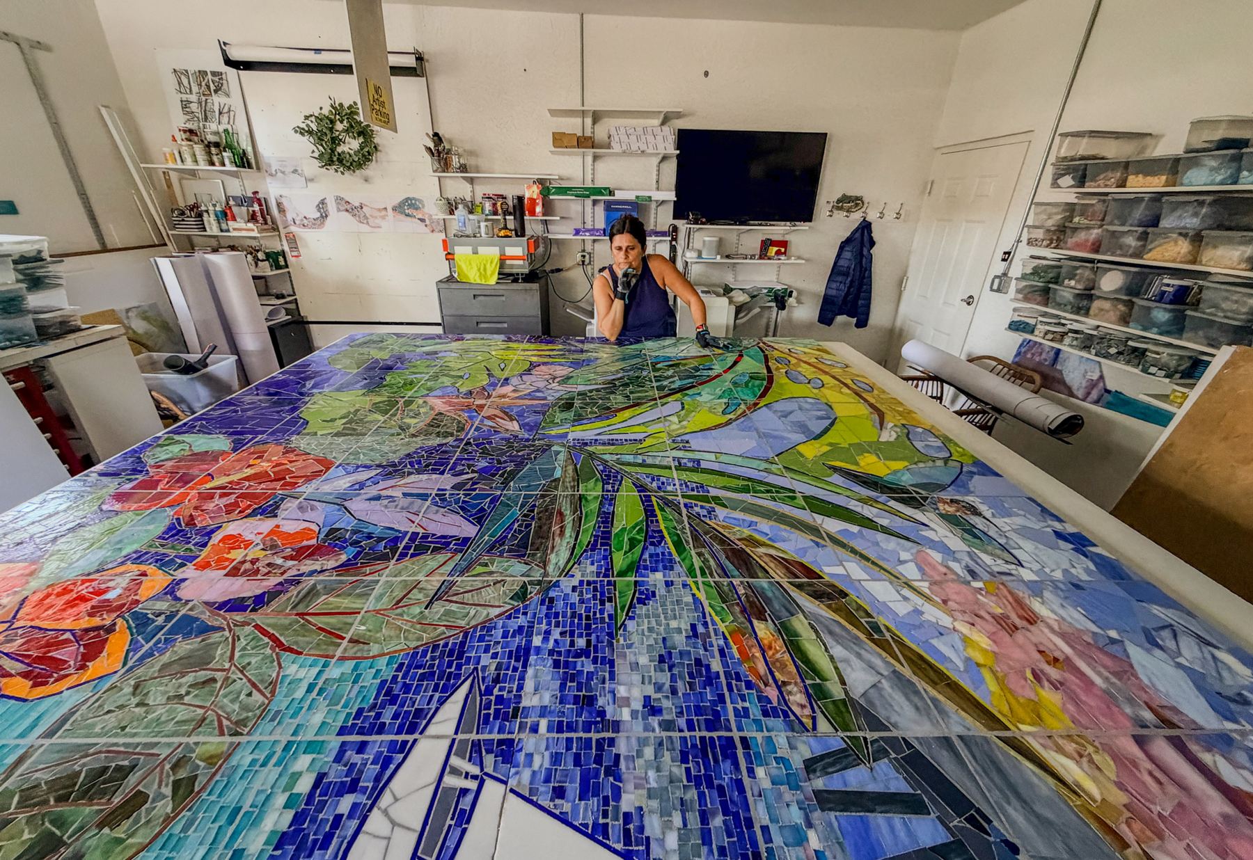 Mila Apperlo, CEO of Apperlo Art, sits behind a monumental handcrafted glass mosaic in the studio, surrounded by neatly organized shelves of materials and tools. The vibrant artwork depicts lush tropical leaves, blooming flowers, and flowing blue water, showcasing the scale, artistry, and craftsmanship behind the creative process.